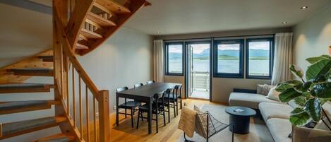 A spacious living area with a wooden staircase, a dining table with chairs, a sofa, and a large window offering a view of the water.