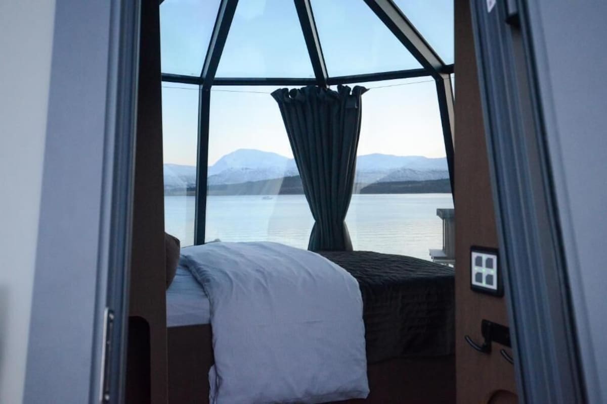 Ein Zimmer mit Bett, einem Vorhangfenster und Blick auf ein Gewässer und Berge.
