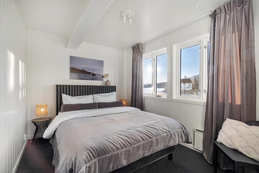 Un dormitorio con cama, mesita de noche, lámpara, ventana con cortinas y un cuadro en la pared.