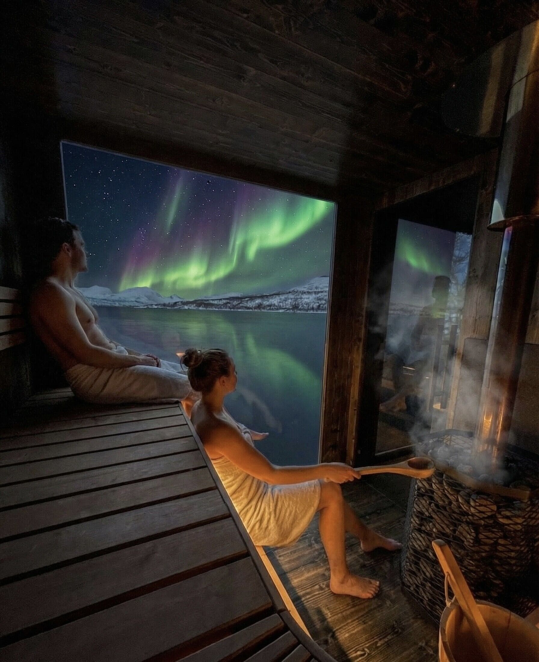 Two people sitting by a window with a view of the northern lights, a wooden deck, and a sauna.
