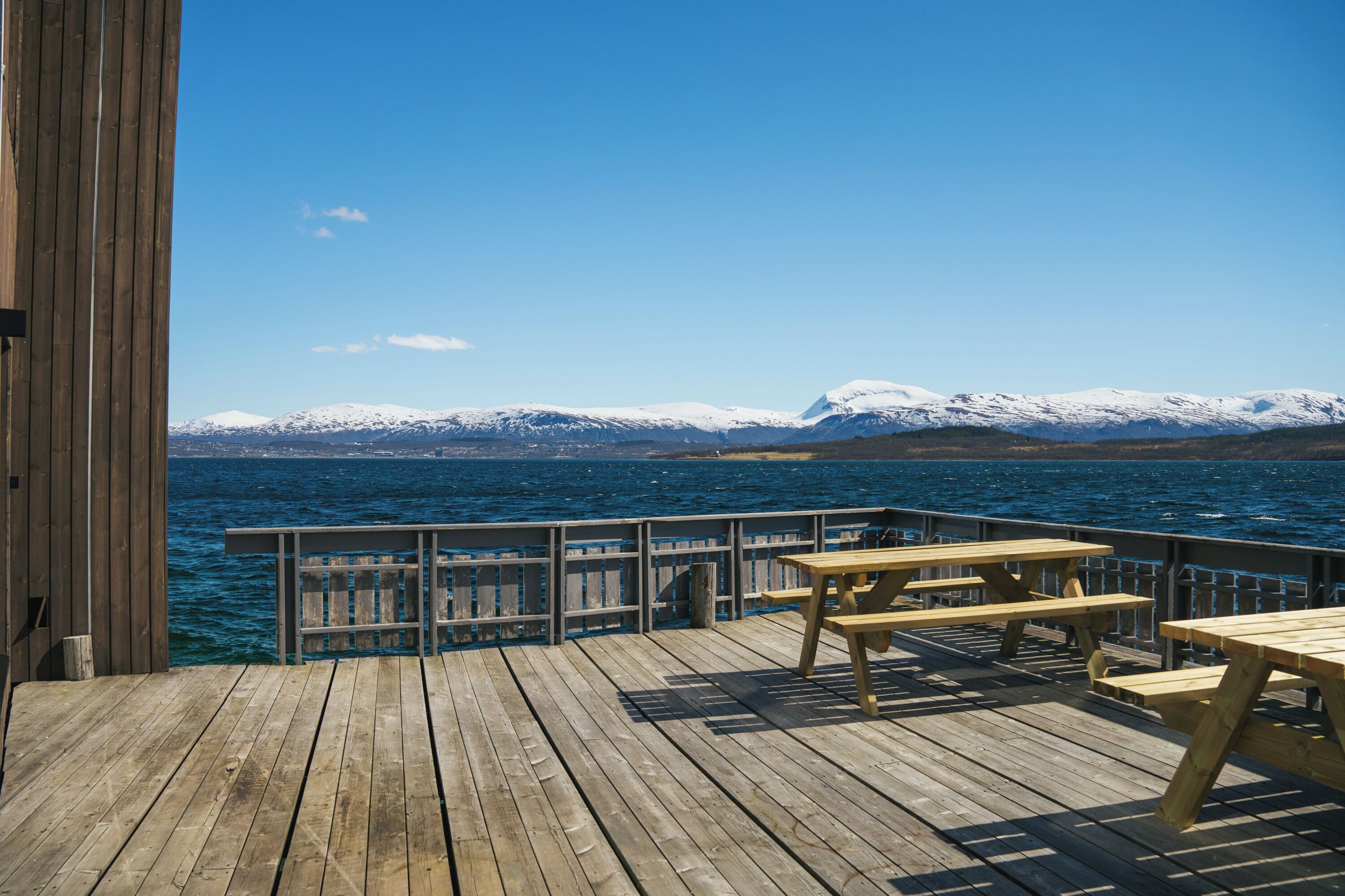 Une terrasse en bois avec des tables de pique-nique, d’où l’on peut admirer un plan d’eau et, au loin, des montagnes enneigées.