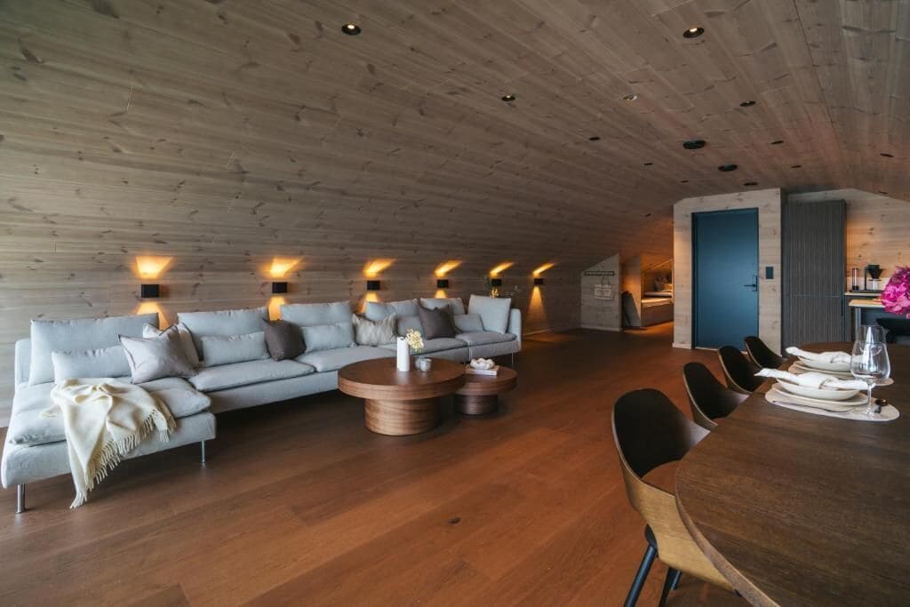 Un interior moderno con techo de madera, un sofá gris, una mesa redonda de madera y un comedor con sillas.