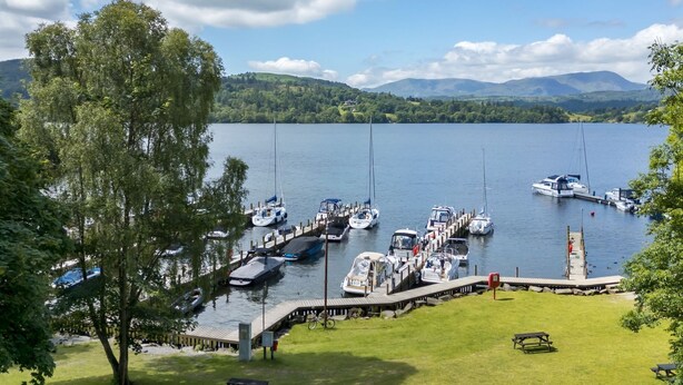 Ein Yachthafen mit vertäuten Booten, eine Rasenfläche mit Bänken und Blick auf einen See mit Bergen im Hintergrund.
