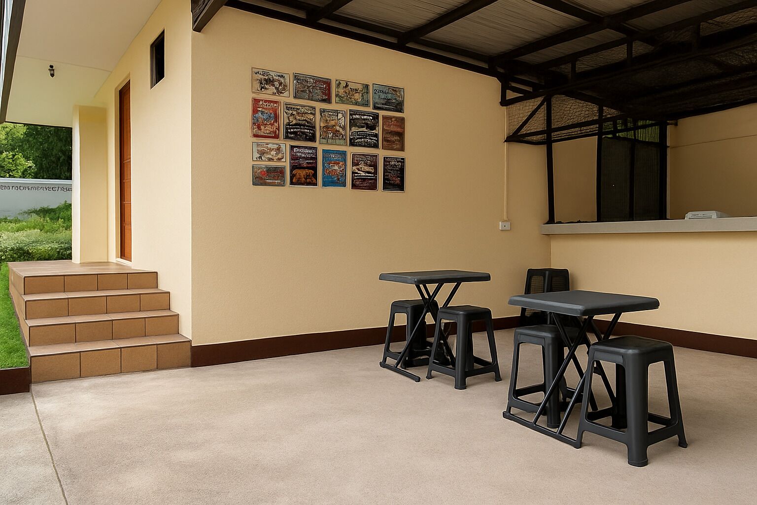 Un espacio al aire libre techado con piso de concreto, paredes color beige y un juego de escaleras que suben. Se observan tres mesas negras y taburetes dispuestos para sentarse.