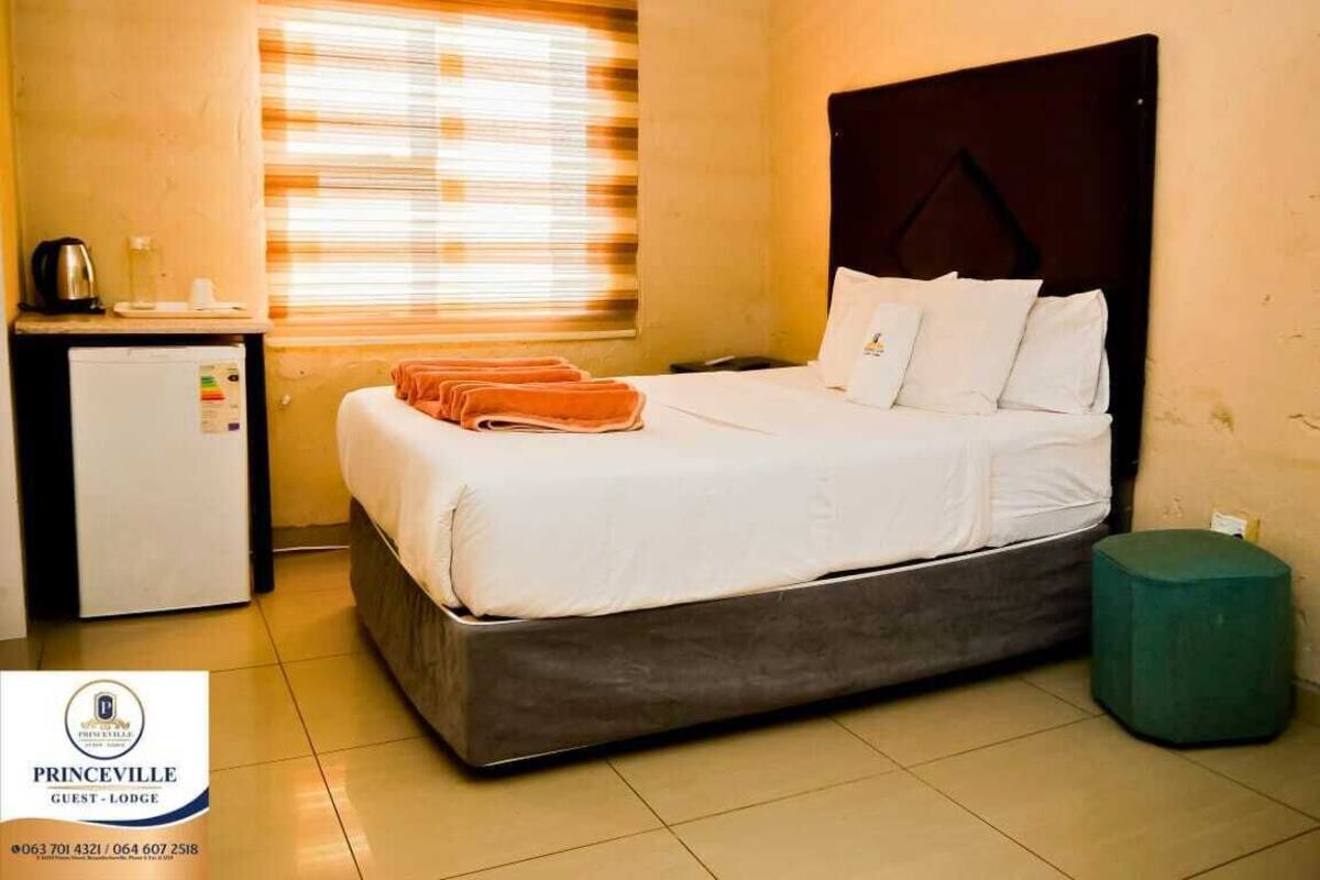 Una habitación de hotel con cama, un escritorio pequeño con hervidor, ventana con persianas y un reposapiés verde.