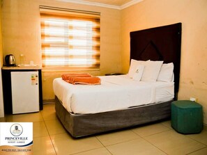 A hotel room with a bed, a small desk with a kettle, a window with blinds, and a green ottoman. - Princeville Guest-Lodge (Soweto)