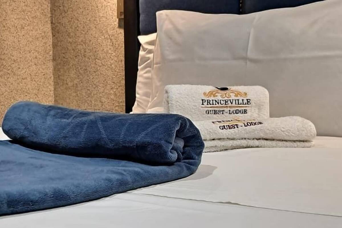 Una cama bien hecha con una manta azul y un cojín blanco con el texto 'PRINCEVILLE GUEST LODGE'.