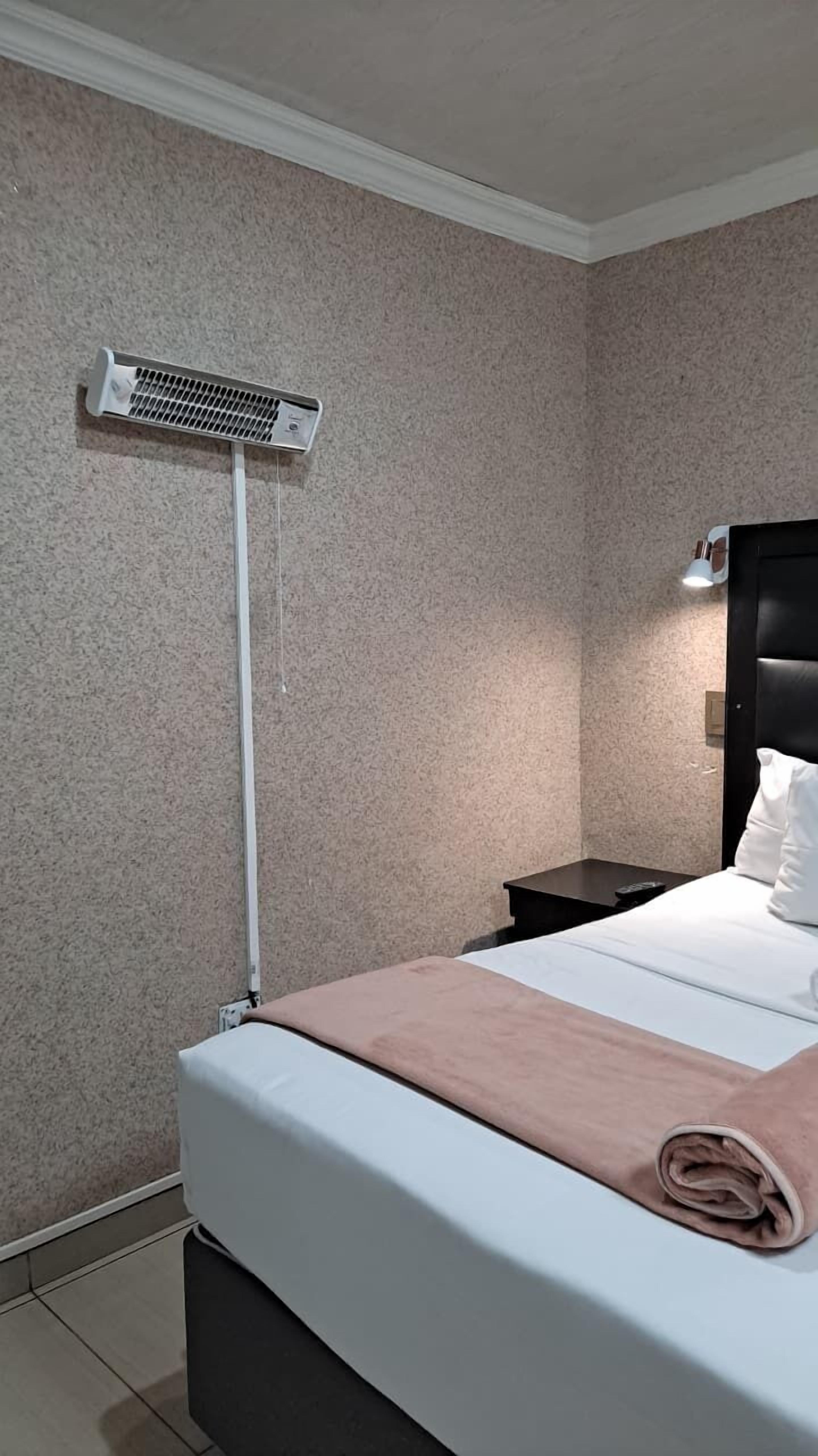 A hotel room with a bed, a bedside table, a wall-mounted heater, and a headboard with a light.