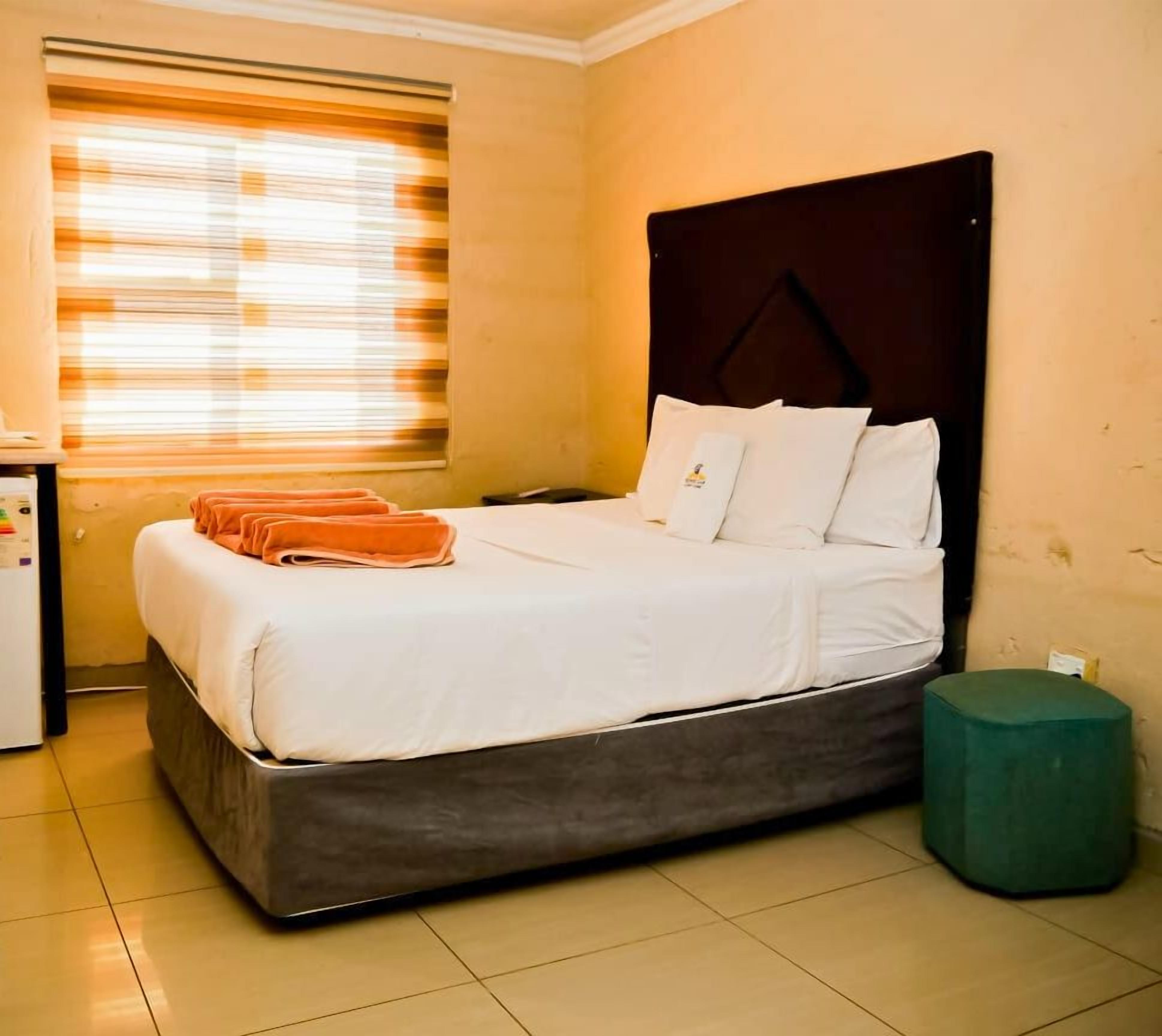Una habitación de hotel con cama, un escritorio pequeño con hervidor, ventana con persianas y un reposapiés verde.