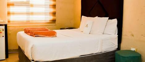 Une chambre d’hôtel avec un lit, un petit bureau avec une bouilloire, une fenêtre avec des stores et un pouf vert.