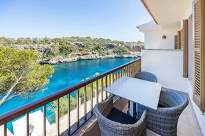 Un balcon avec une table et des chaises donnant sur une zone côtière, avec une eau d’un bleu limpide et un paysage rocheux.