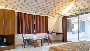 A hotel room with a bed, a desk, two chairs, a small table, a minibar, and a view of the outside. - Road to Heaven Resort (Bhuj)