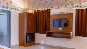 A bedroom with a built-in wardrobe, a small refrigerator, a mounted TV, and a bed with a patterned bedspread. - Road to Heaven Resort (Bhuj)