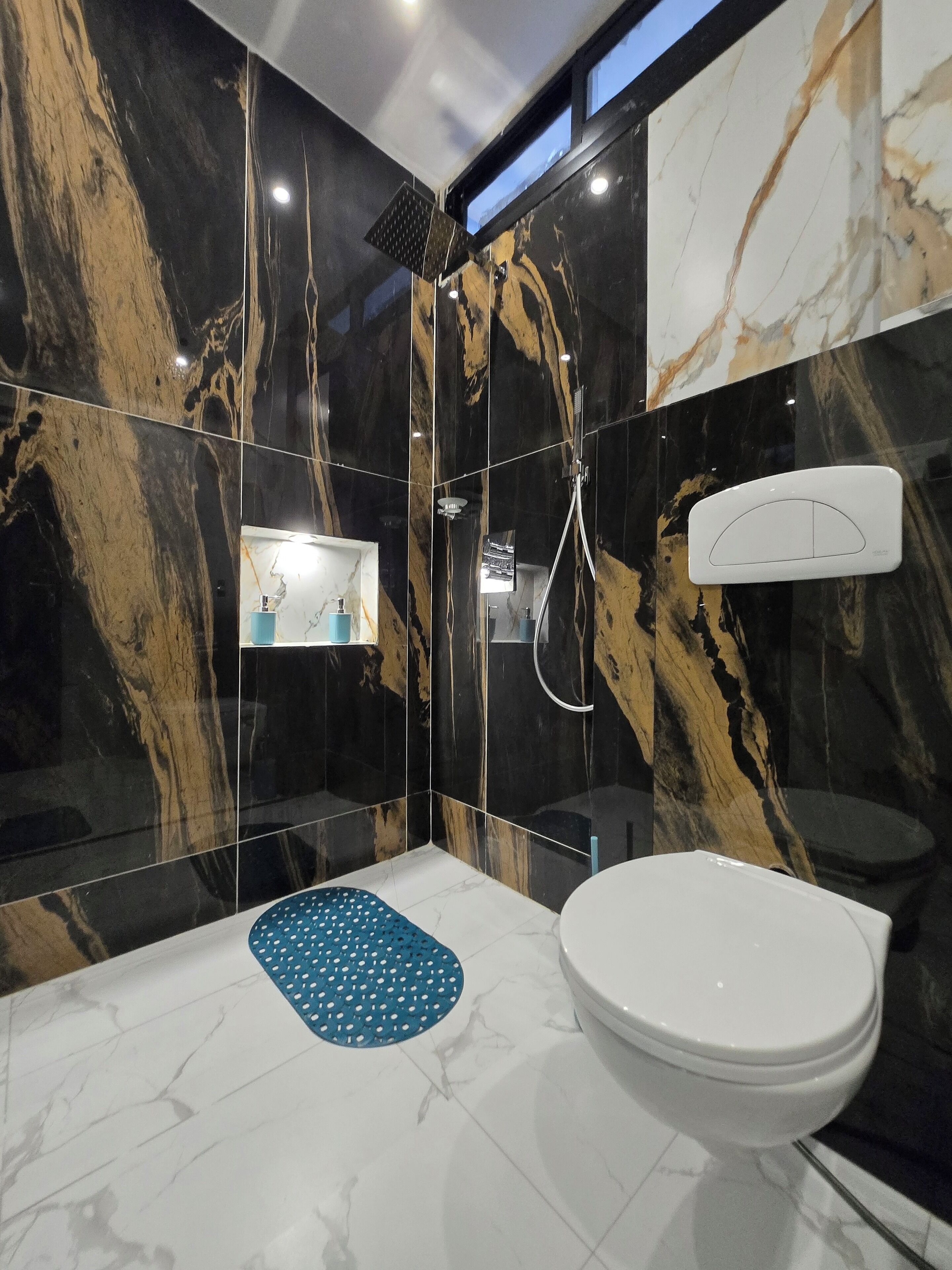 A modern bathroom with a marble wall, a white toilet, a blue mat, and a shower.