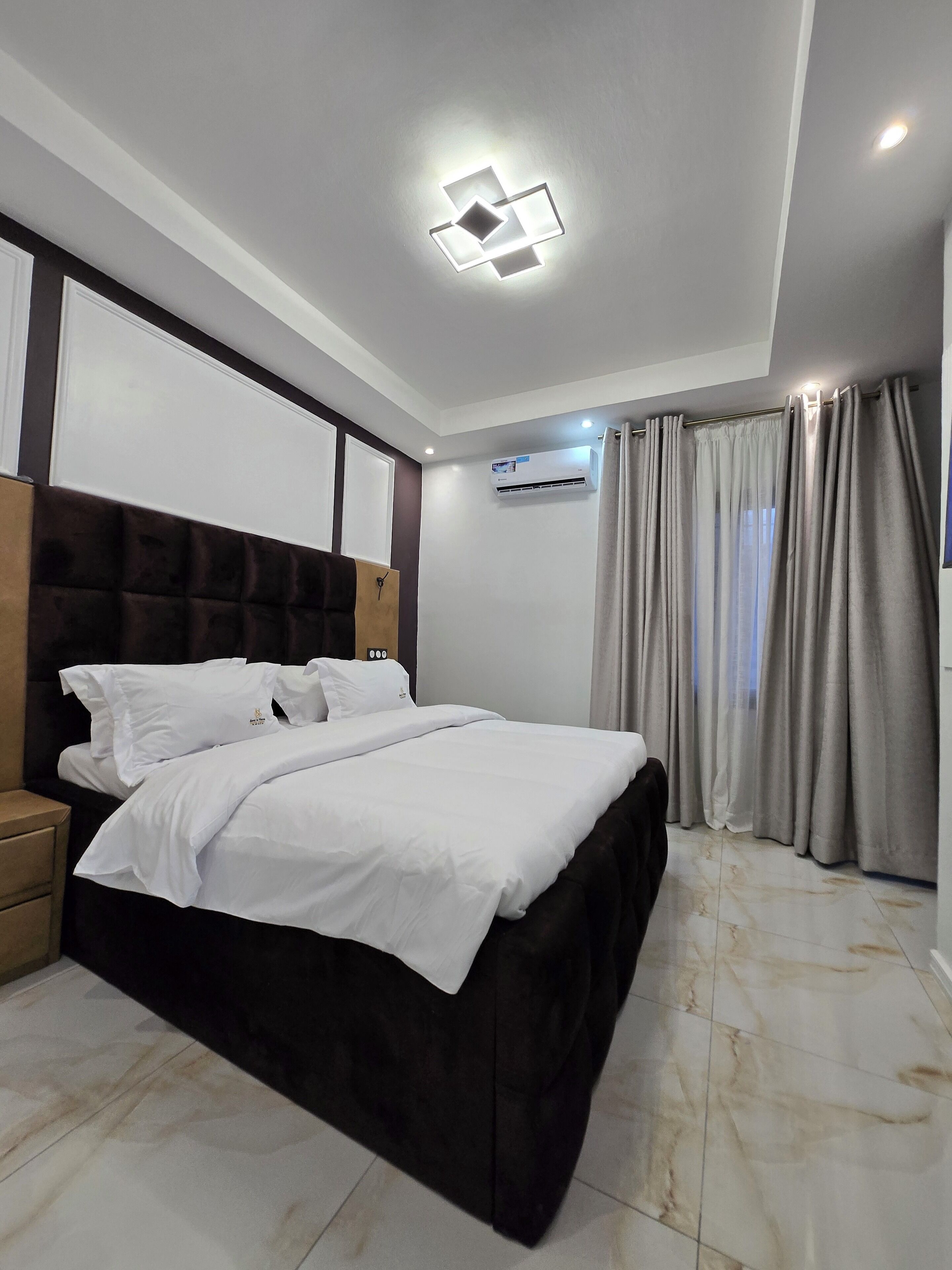 Un dormitorio moderno con una cama grande, un cabecero, un equipo de aire acondicionado y una ventana con cortinas.