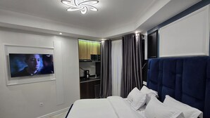 Una camera d'albergo moderna con un letto grande, una televisione montata a parete, un angolo cottura piccolo e un bagno visibile attraverso un pannello di vetro.