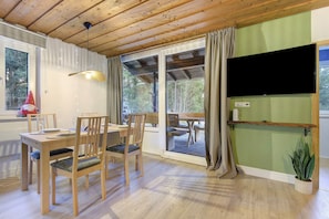 A dining area with wooden furniture, a TV mounted on the wall, and a large window with curtains. - Holiday Home 'Am Neubäuer See' with Private Terrace, Shared Garden, and Wi-Fi (Roding)