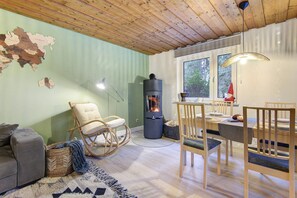 A cozy room with a wooden ceiling, a dining table with chairs, a sofa, a rocking chair, a fireplace, and a large window. - Holiday Home 'Am Neubäuer See' with Private Terrace, Shared Garden, and Wi-Fi (Roding)