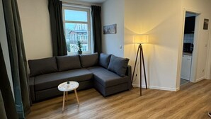 Un salon moderne avec un canapé d’angle, une petite table ronde, un lampadaire et une vue sur un immeuble par la fenêtre.
