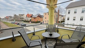 Un balcon avec une table et des chaises, donnant sur une rue bordée d’immeubles et de voitures garées.