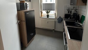 Une cuisine compacte équipée d’un four encastré, d’un micro-ondes et d’un évier. On y trouve une fenêtre avec des stores, un radiateur et un pot de fleurs posé sur le rebord de la fenêtre.