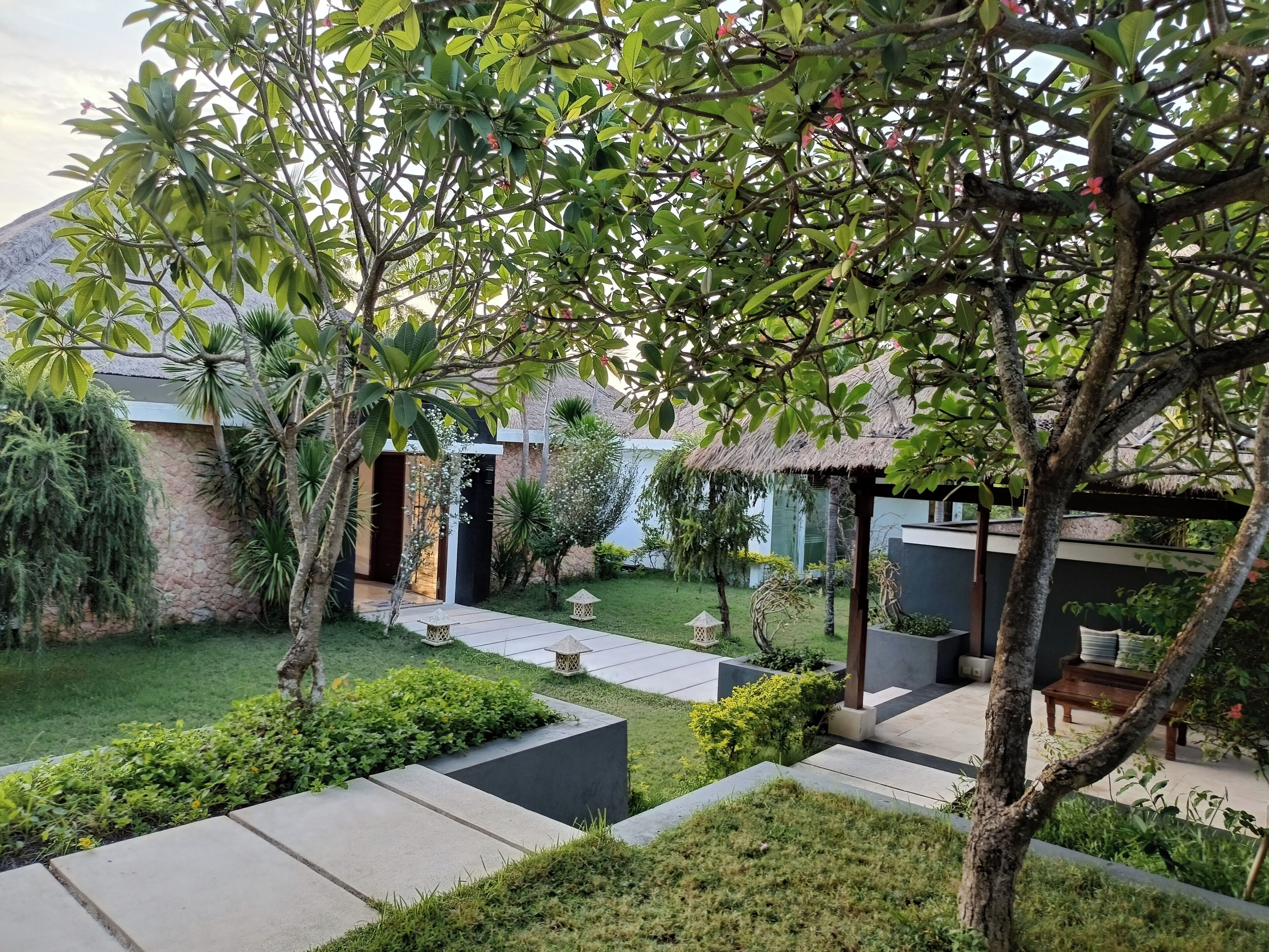 A landscaped garden with a thatched-roof structure, a paved pathway, and various plants.