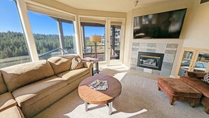 A living room with a sofa, coffee table, fireplace, TV, and large windows overlooking a forest. - River Ridge 324AB Ground Floor Condo in Bend, OR 2 Bd, 2 Ba (Bend)
