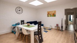 A modern living room with a dining area, featuring a white table, chairs, a blue sofa, a clock on the wall, and a painting.