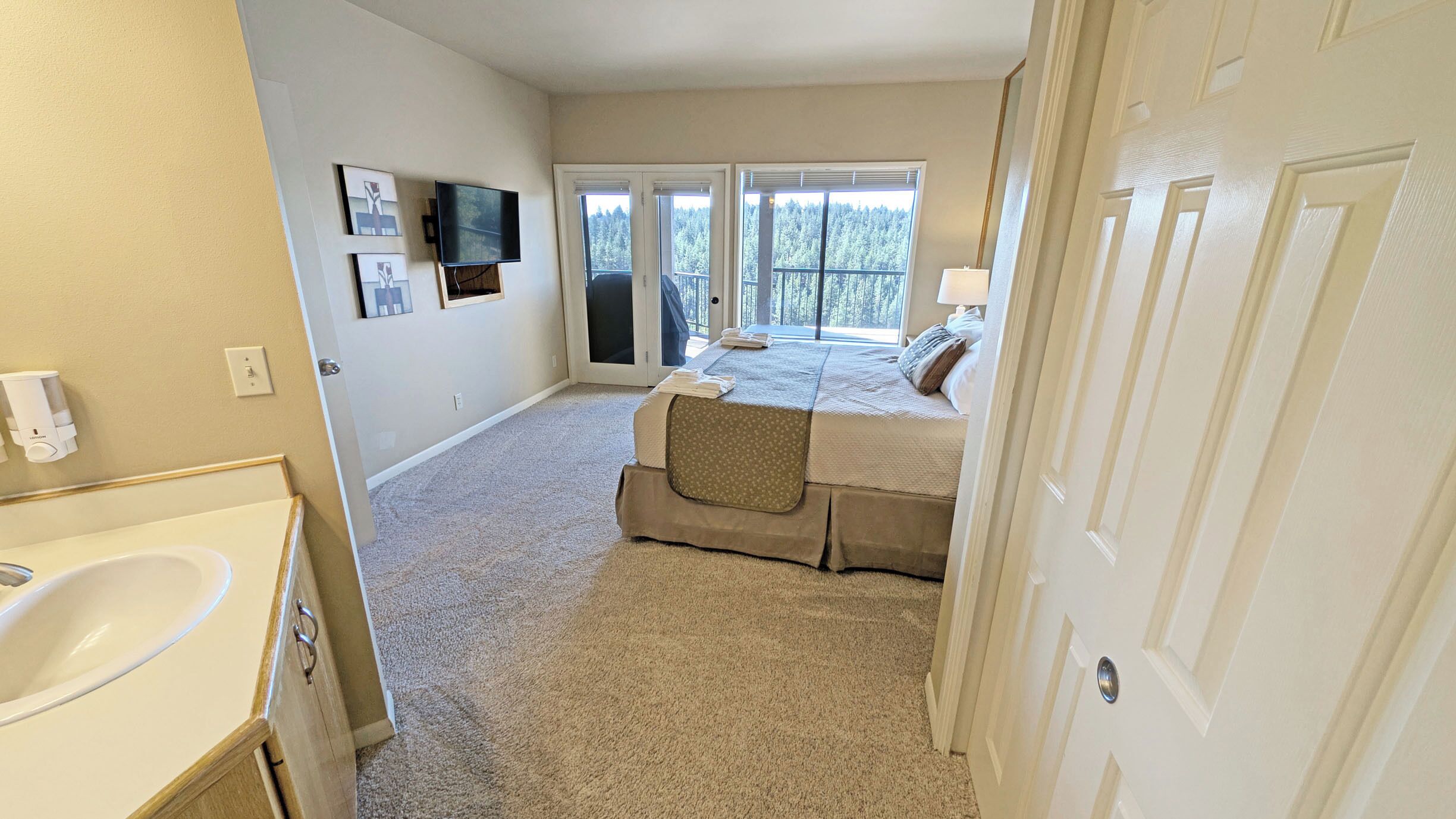 A bedroom with a large window, a bed, a TV, and a bathroom with a sink.