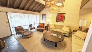 Un salon chaleureux avec un canapé, un fauteuil, un pouf et une table basse. On y trouve des lampes en osier, un tableau encadré et une lampe sur une table d’appoint.