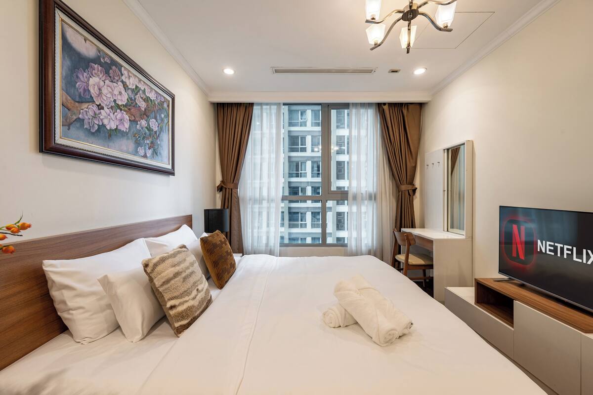 A hotel room with a large bed, a painting on the wall, a TV with the Netflix logo, and a window with curtains.