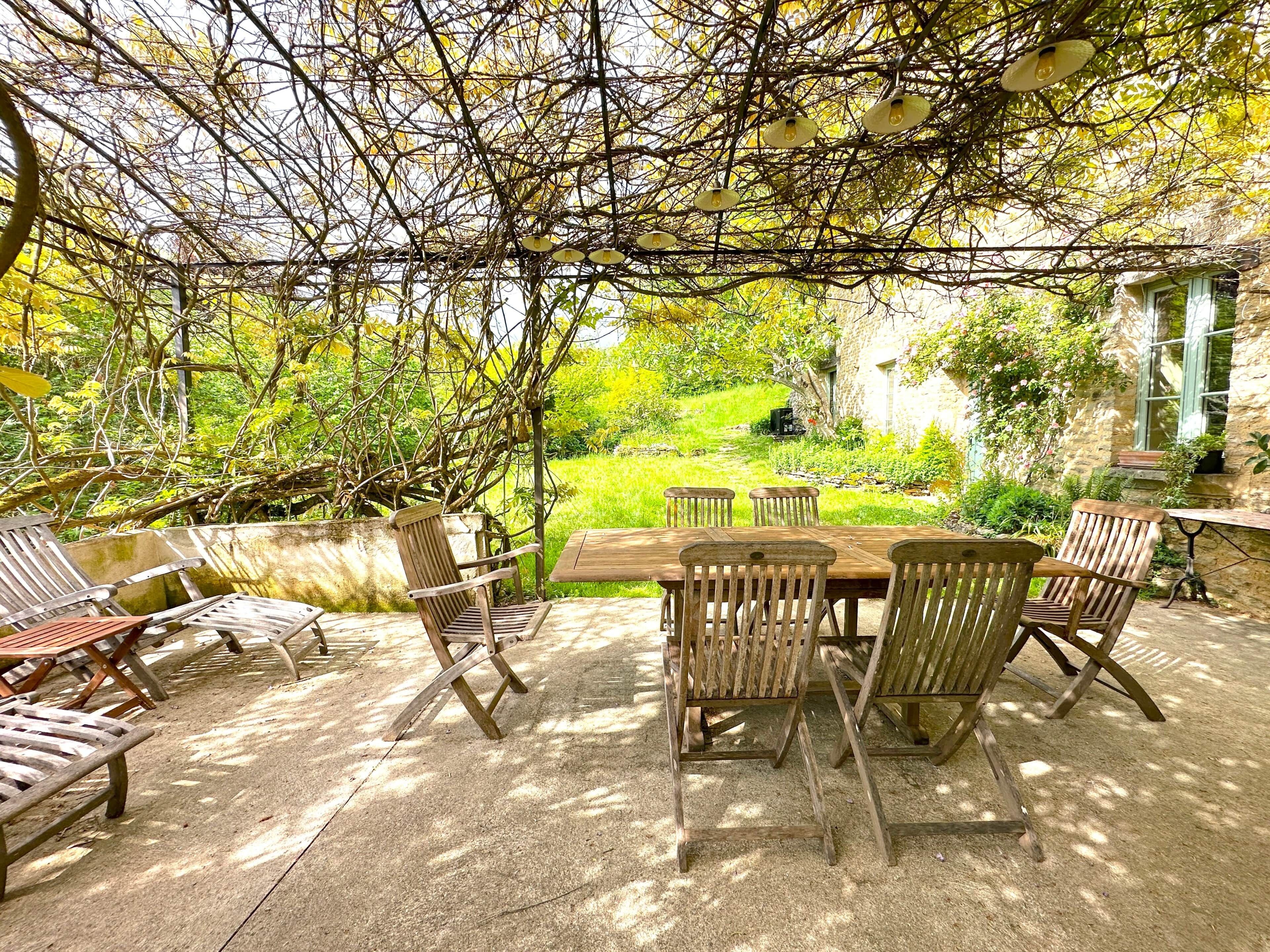 Pátio externo com mesa e cadeiras de madeira, cercado por vegetação e uma parede de pedra.