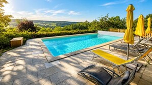 A swimming pool with a view of the countryside, surrounded by greenery and equipped with sun loungers and umbrellas.