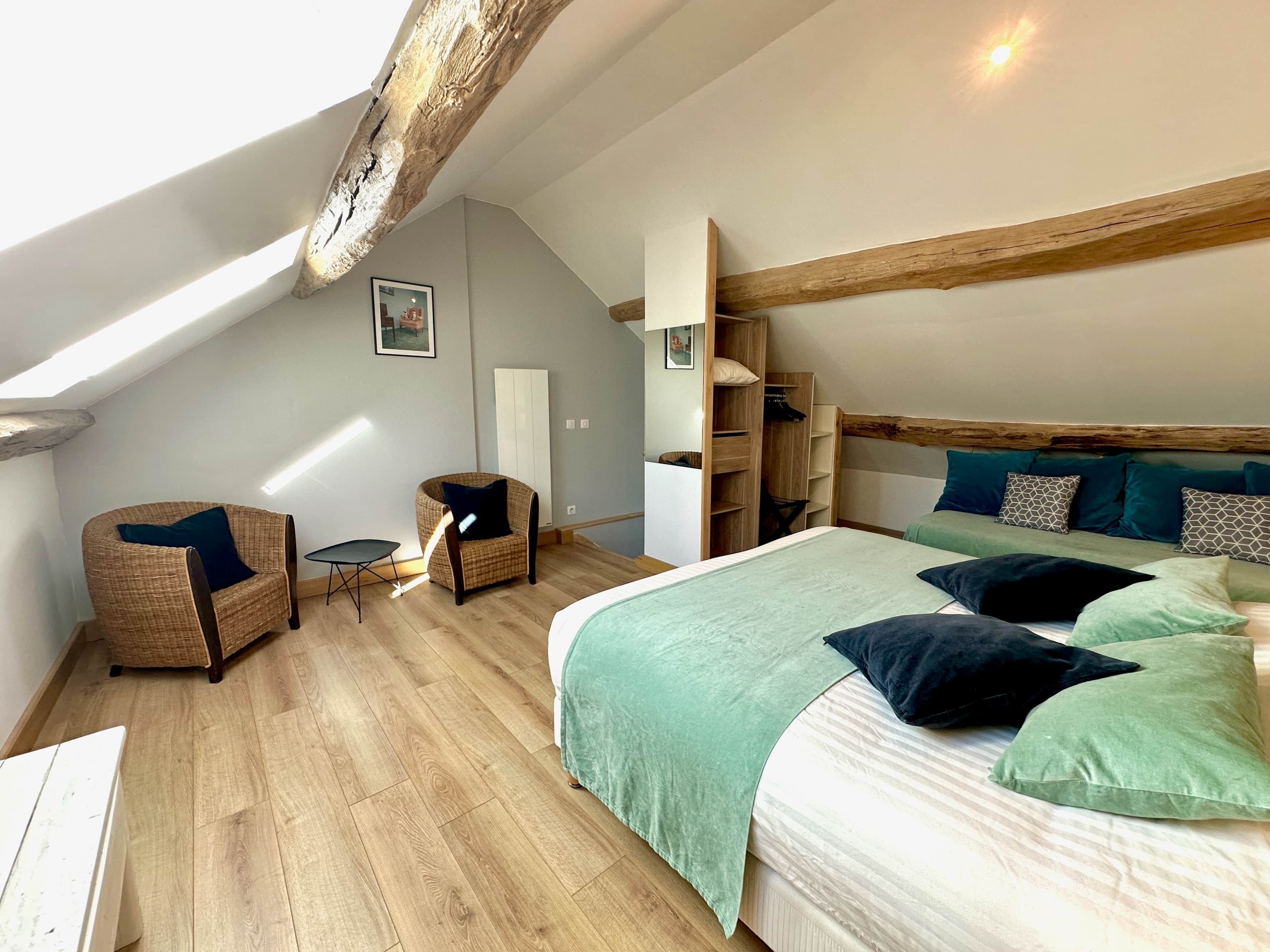 Un dormitorio con techo inclinado, vigas de madera, una cama con almohadas verdes y negras, una silla de mimbre y una mesita.