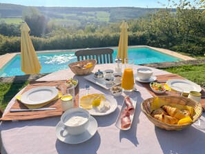 A breakfast table with various food items and a glass of orange juice, overlooking a swimming pool.