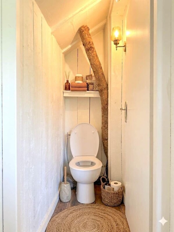A narrow bathroom with a white toilet, a wooden tree trunk, a shelf with toiletries, and a wall-mounted light fixture.