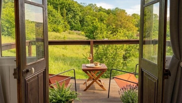 An open door leading to a balcony with a table and chairs, surrounded by greenery and a clear sky.
