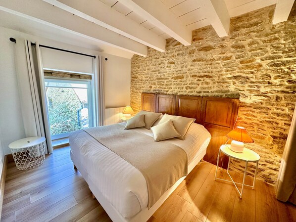 A bedroom with a stone wall, a large bed with pillows, a nightstand with a lamp, and a window with curtains. - La Grange 4/5 personnes (Montoillot)