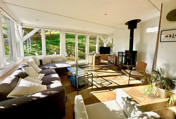 Un salon lumineux, avec un canapé, une table basse en verre, un téléviseur, une cheminée et une vue sur la verdure à l’extérieur.