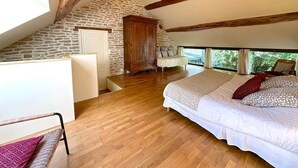 A bedroom with a sloped ceiling, wooden beams, a stone wall, a bed with patterned bedding, and a view of greenery outside. - The Fleurières (Montoillot)