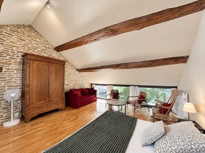Une pièce avec un mur en pierre, des poutres en bois, un canapé rouge, une table à manger avec des chaises et un lit agrémenté d’oreillers à motifs.