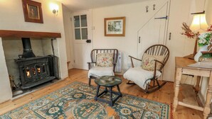 Une pièce chaleureuse avec un poêle à bois, deux fauteuils à bascule, un tapis à motifs et une table ornée d’un vase de fleurs.