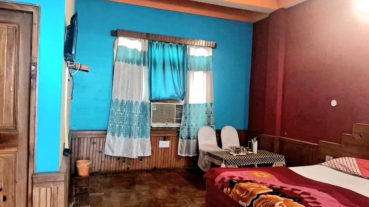 Ein Zimmer mit einem Bett, einem kleinen Tisch, zwei Stühlen, einem Fenster mit Vorhängen und einer Wand-Klimaanlage.
