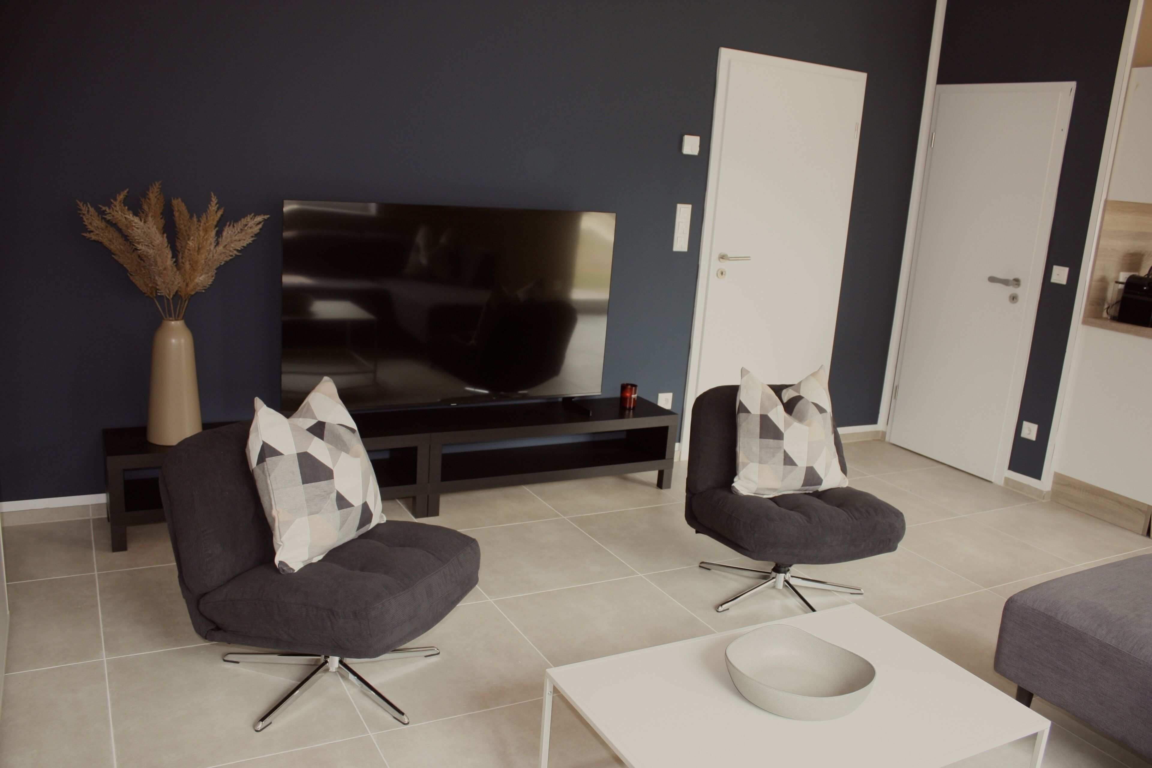 A modern living room with a flat-screen TV, two grey armchairs, a white coffee table, and a vase with dried plants.