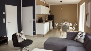 A modern living room with a sofa and armchair, a dining area with a table and chairs, and a kitchenette with cabinets and appliances. - Stylish living - job & vacation (Soltau)