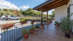 A covered outdoor area with potted plants, a metal fence, and a building with a sloped roof.