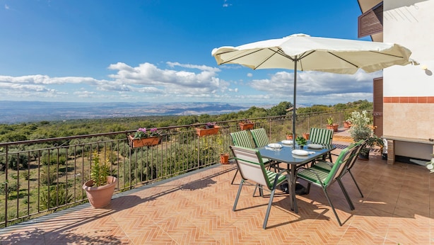 An outdoor patio with a table and chairs, a white umbrella, and a view of the landscape.