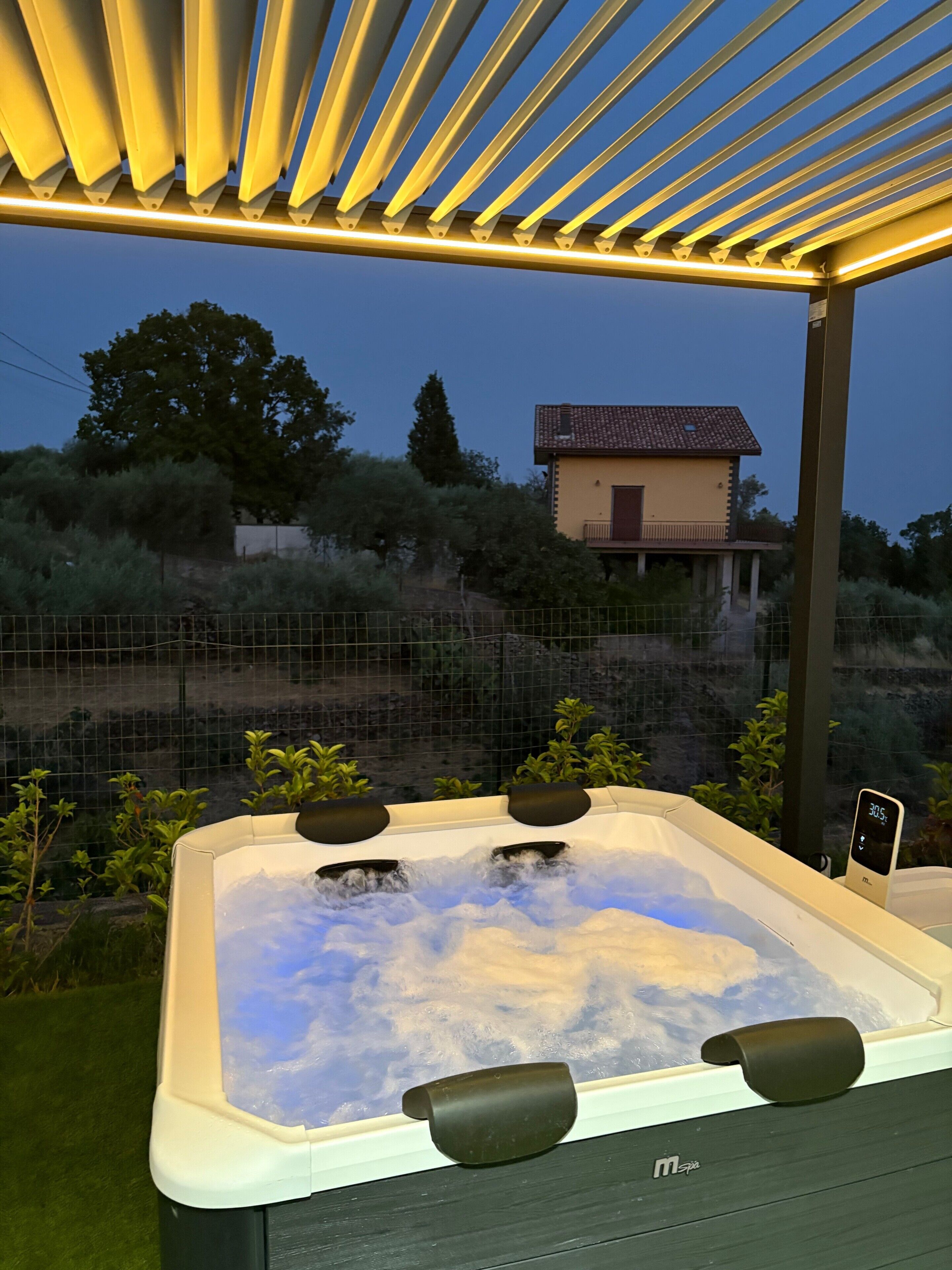 A hot tub with three jets, surrounded by a pergola with overhead lighting, set against a backdrop of a house and trees.