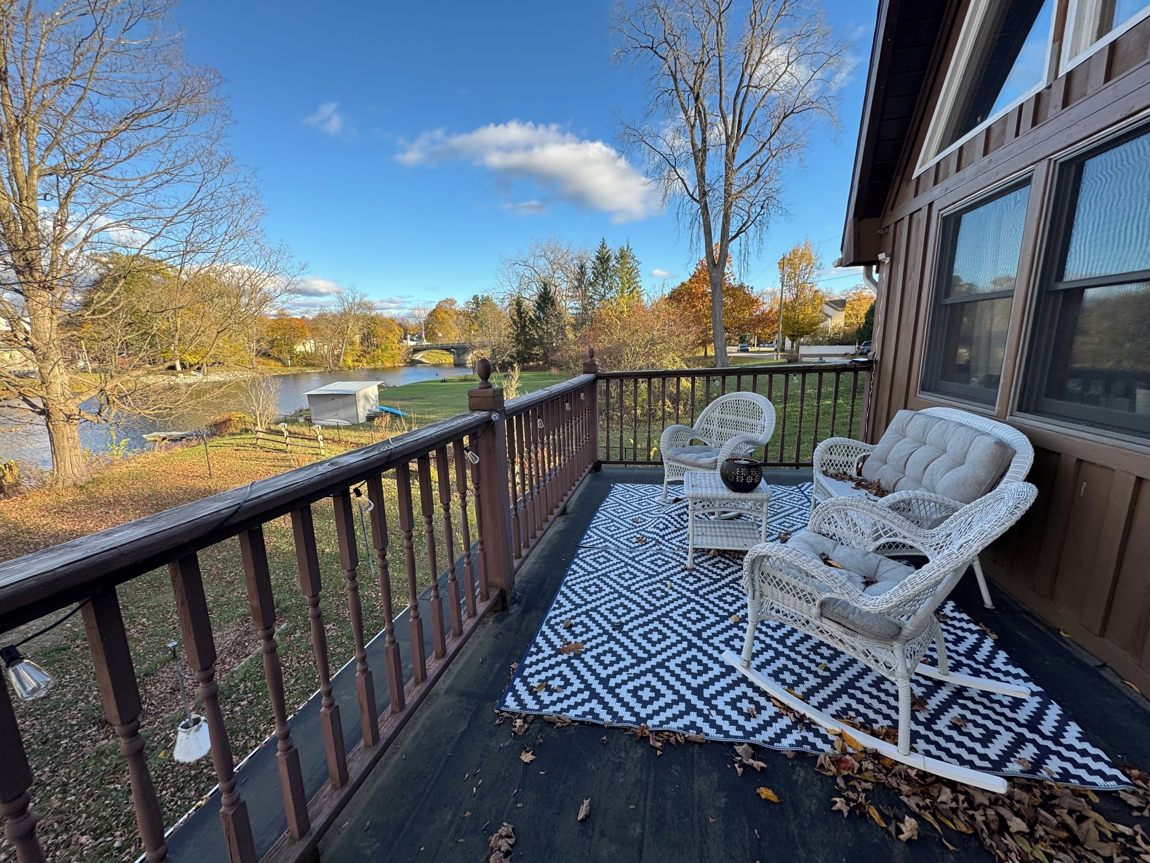 A balcony with wicker furniture and a patterned rug, overlooking a river and autumn trees.