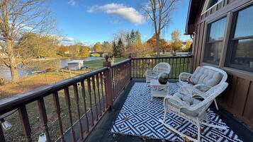 A balcony with wicker furniture and a patterned rug, overlooking a river and autumn trees.